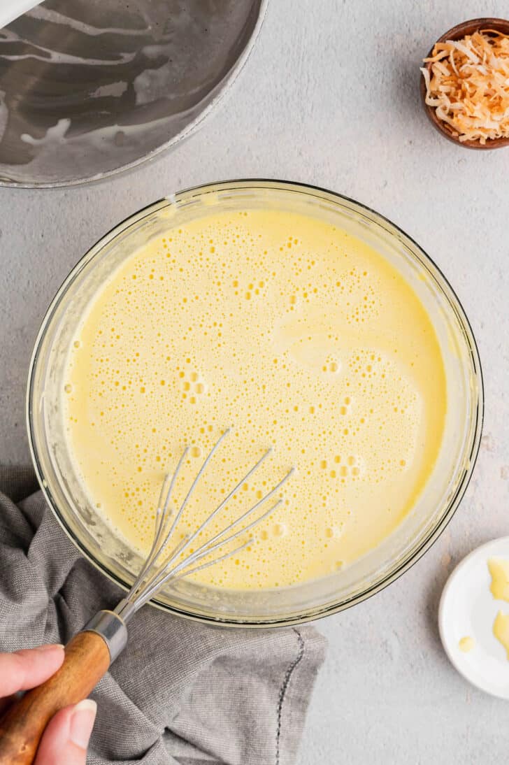 A glass bowl filled with a light yellow bubbly liquid, with a whisk stirring the liquid.