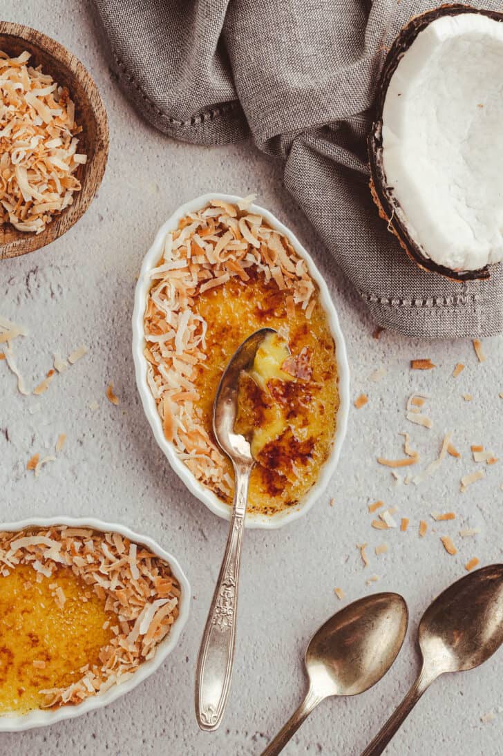 A white oval ceramic dish filled with a custard with a torched sugar top, garnished with shredded coconut, with a metal spoon digging in to the dessert.