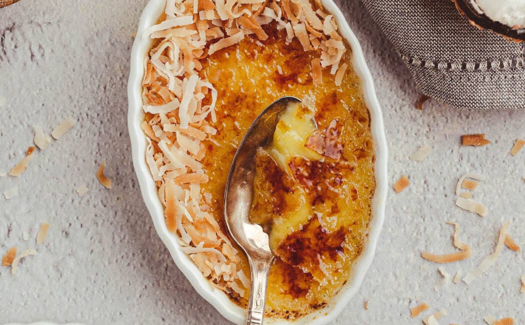 A white oval ceramic dish filled with a custard with a torched sugar top, garnished with shredded coconut, with a metal spoon digging in to the dessert.