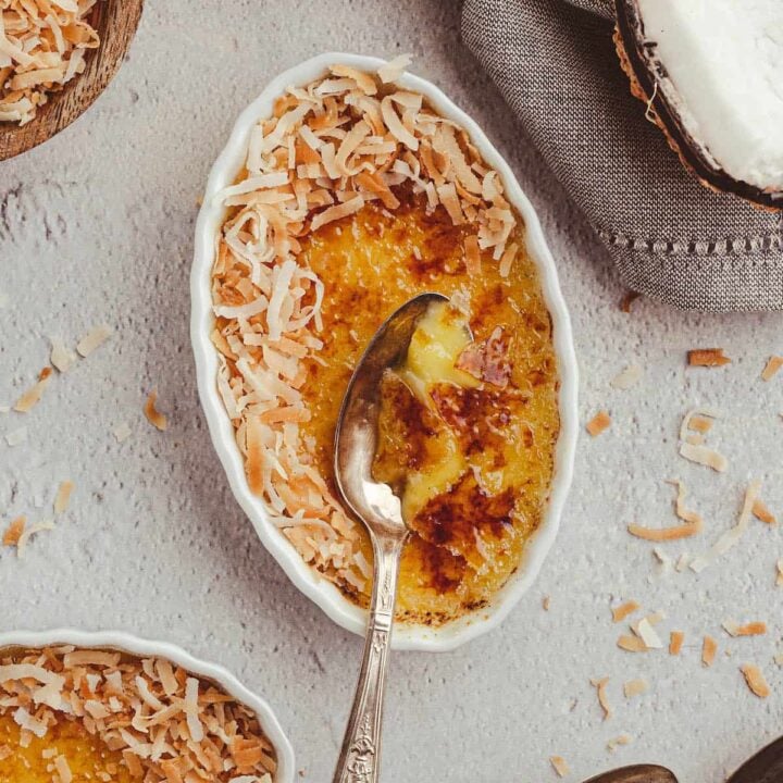 A white oval ceramic dish filled with a custard with a torched sugar top, garnished with shredded coconut, with a metal spoon digging in to the dessert.
