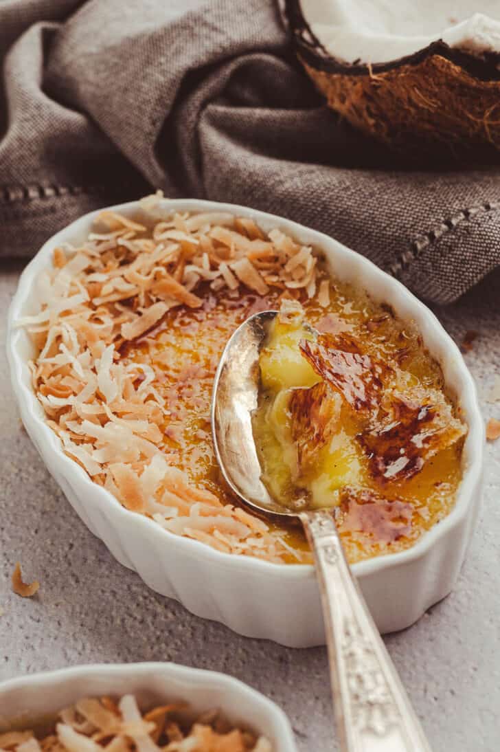 An oval shaped white ceramic dish filled with coconut creme brulee, garnished with toasted coconut, with a spoon digging into the custard.