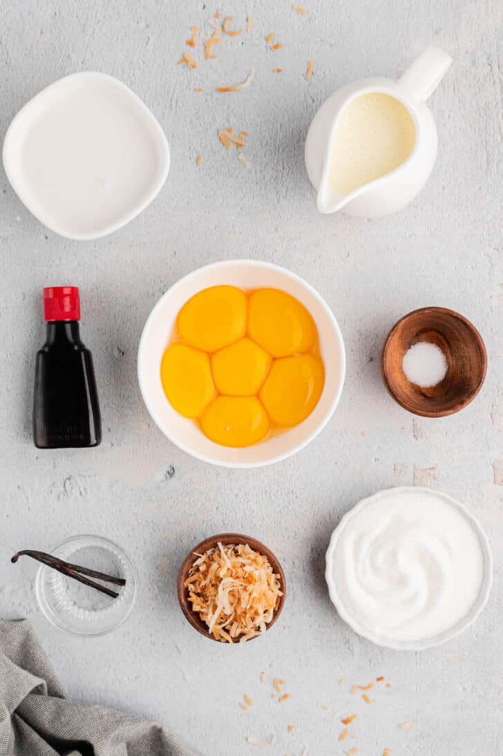 A light surface with all of the ingredients for coconut creme brulee displayed in small bowls, including egg yolks, sugar, salt, toasted coconut, vanilla beans and extract.