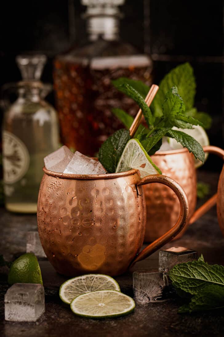 Two hammered copper mugs filled with a Kentucky mule cocktail, garnished with lime wedges and mint sprigs.