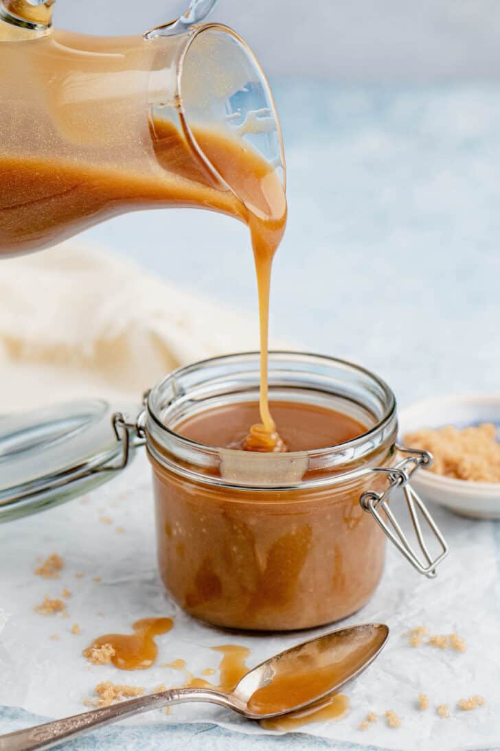 A glass pitcher pouring microwave caramel sauce into a small lidded jar.