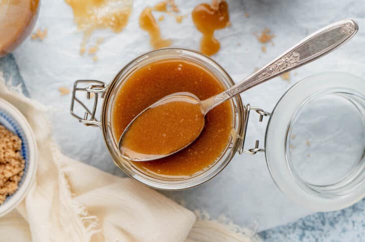 A small lidded glass jar, open, filled with caramel sauce, with a spoon scooping some sauce out.