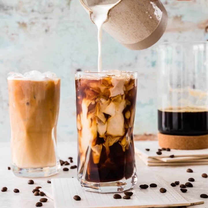 A large glass of iced coffee with cream from a pitcher being poured into it. The scene is garnished with napkins, a pitcher of coffee, and coffee beans.