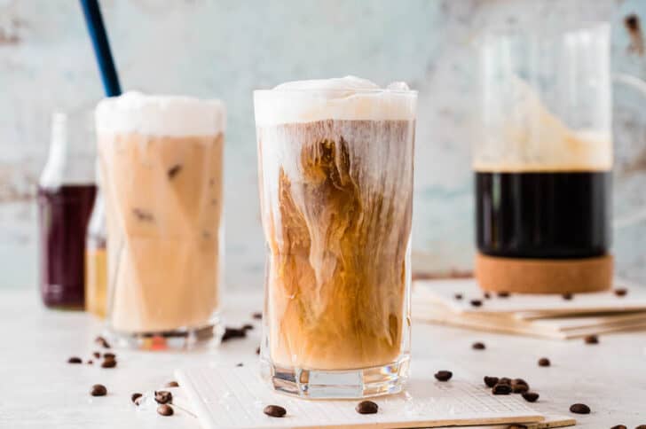 A large glass of iced coffee with cream recently poured into it, with a pitcher of coffee in the background.