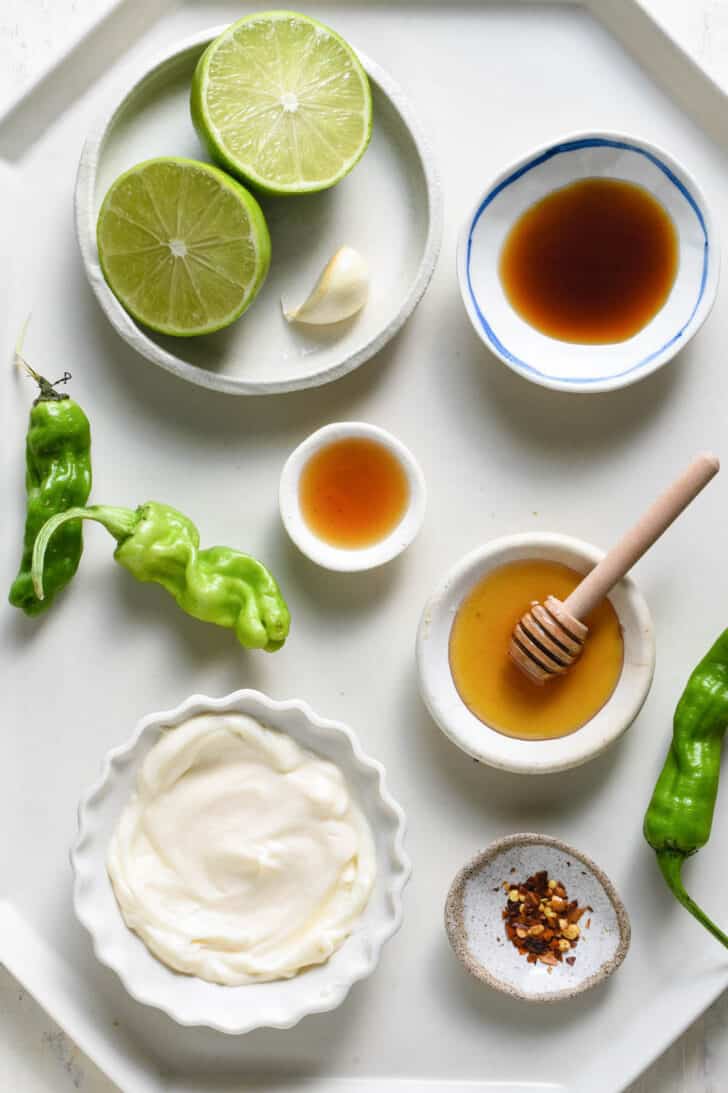 All of the ingredients needed for creamy condiment sauce, including a lime, garlic clove, soy sauce, fish sauce, honey, mayonnaise and red pepper flakes, on a white geometric tray.
