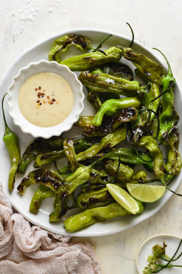 Round white plate filled with blistered green vegetables with stems and a small fluted white bowl of creamy light orange sauce with red pepper flakes sprinkled on top.