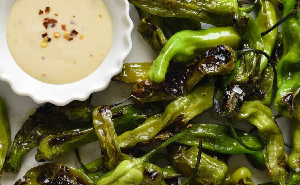 Round white plate filled with blistered shishito peppers and a small fluted white bowl of creamy light orange sauce with red pepper flakes sprinkled on top.