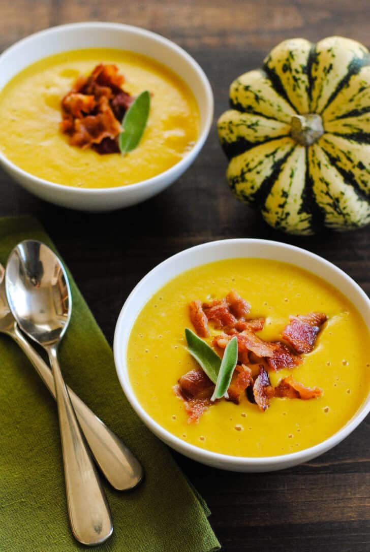 Two small white bowls filled with sweet dumpling squash soup garnished with chopped bacon and sage leaves, with a green napkin, two spoons and a whole dumpling squash nearby.