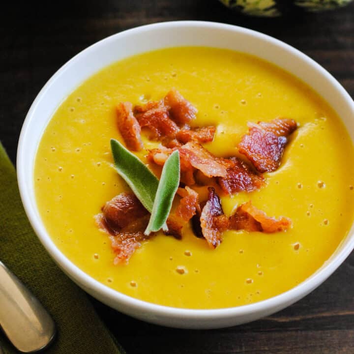 A small white bowl filled with sweet dumpling squash soup garnished with chopped bacon and sage leaves, on a dark surface.