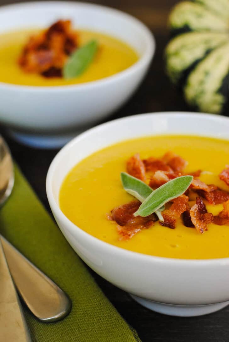 Two small white bowls filled with sweet dumpling squash soup garnished with chopped bacon and sage leaves, with a green napkin, two spoons and a whole dumpling squash nearby.