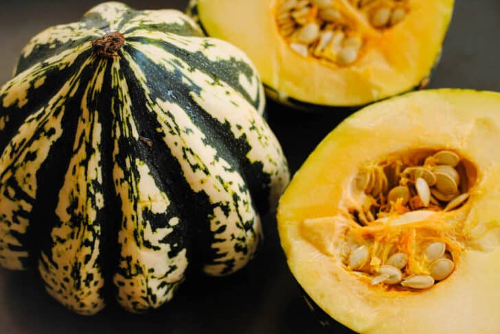 Two dumpling squashes on a dark surface, one whole and one cut in half exposing the flesh and seeds.