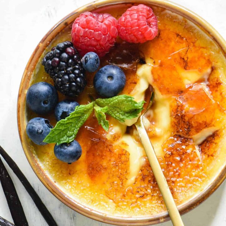 Shallow dish of vanilla bean creme brulee garnished with berries and mint, with a gold spoon digging in, with vanilla beans alongside.