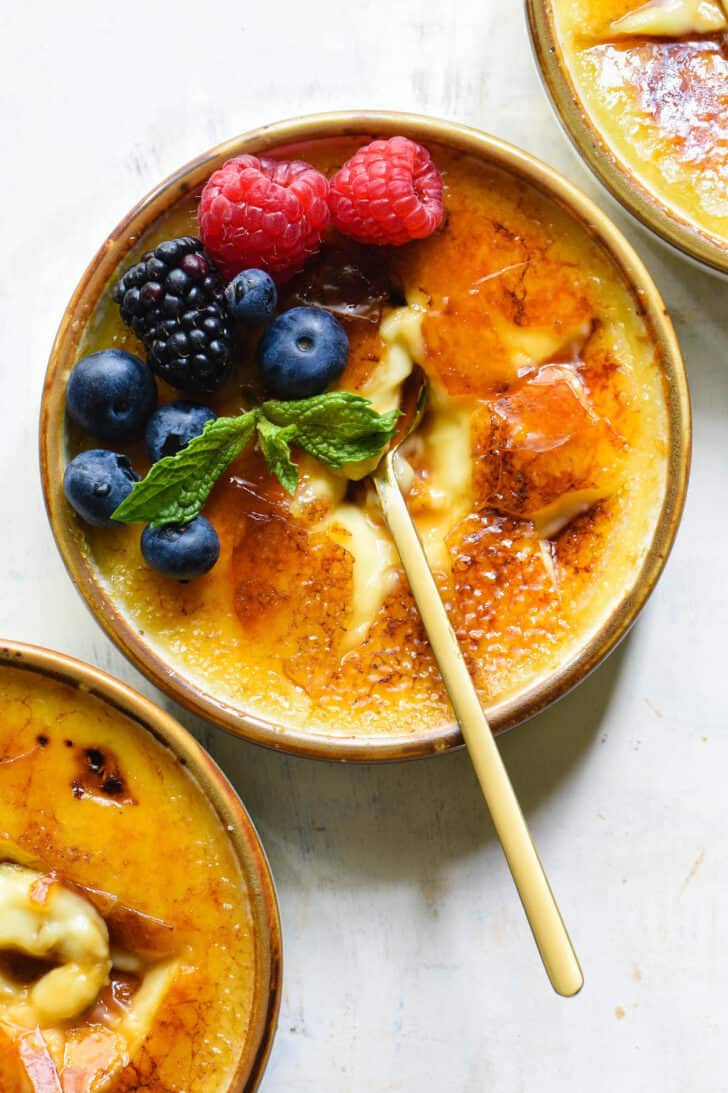 Closeup on a gold-rimmed ramekin of vanilla creme brulee garnished with blueberries, raspberries, a blackberry and a mint sprig, with a gold spoon breaking through the torched sugar.
