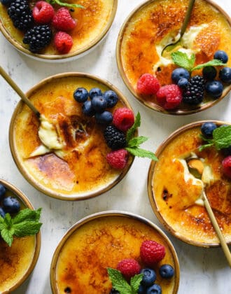 Six dishes of vanilla bean creme brulee garnished with berries and mint sprigs, on a light surface, with gold spoons digging into three of the dishes.