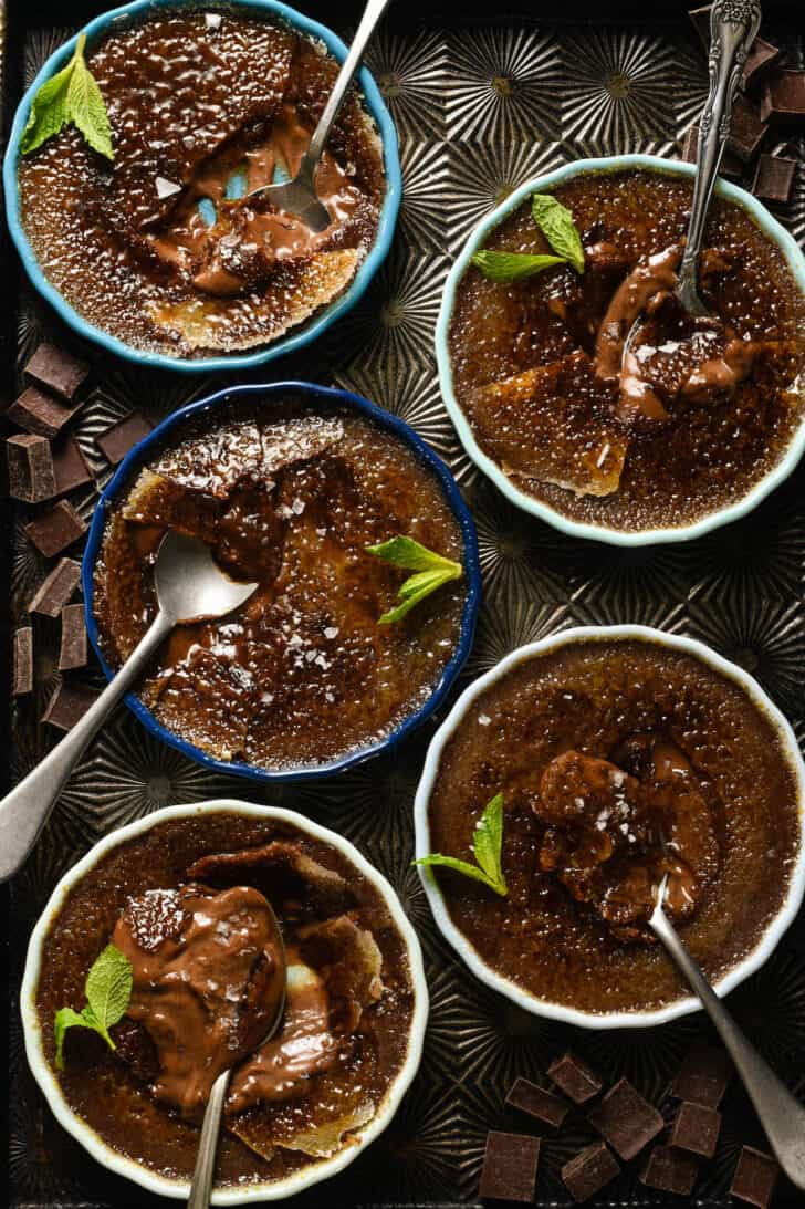 Five shallow dishes of chocolate custard garnished with mint sprigs and sea salt, with spoons digging into them, on a textured baking pan.