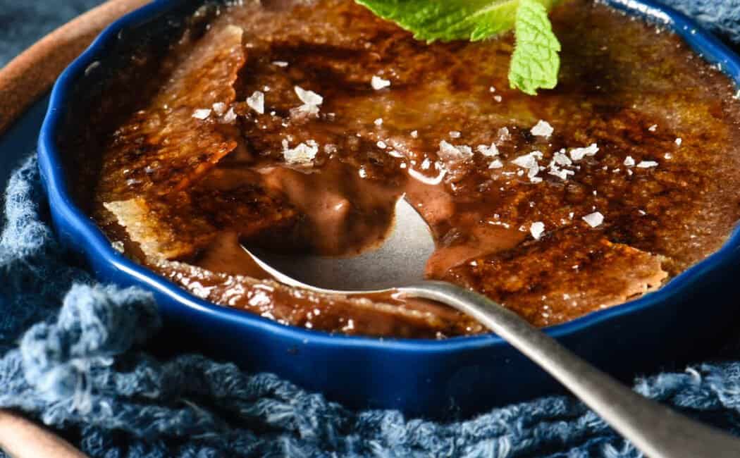 A shallow dark blue ramekin filled with chocolate creme brulee, garnished with a mint sprig and sea salt flakes, with a spoon digging into it.