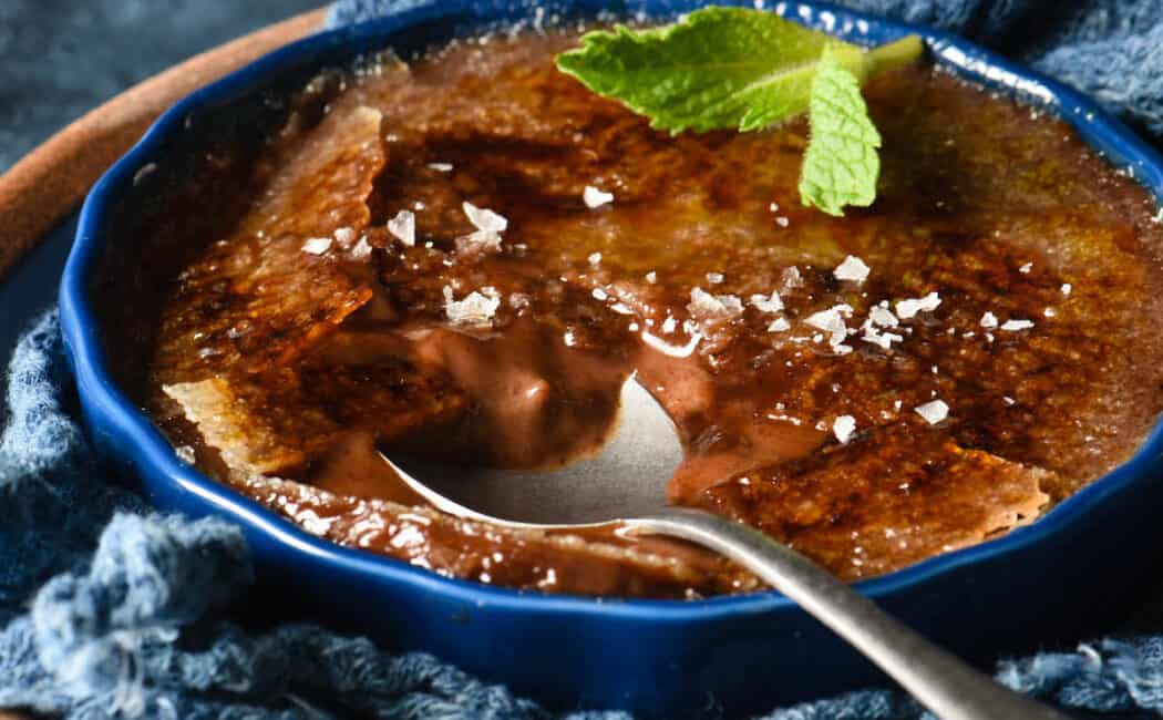 A shallow dark blue ramekin filled with chocolate creme brulee, garnished with a mint sprig and sea salt flakes, with a spoon digging into it.