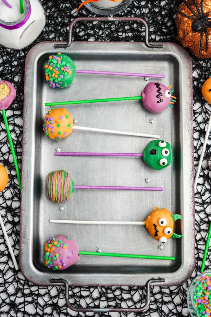 Pumpkin cake pops coated in colored candy coating and decorated with candy eyeballs and sprinkles, on a silver serving tray.
