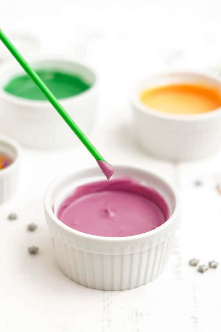 A white ramekin filled with melted purple candy coating, with a green lollipop stick just having been dipped into it.