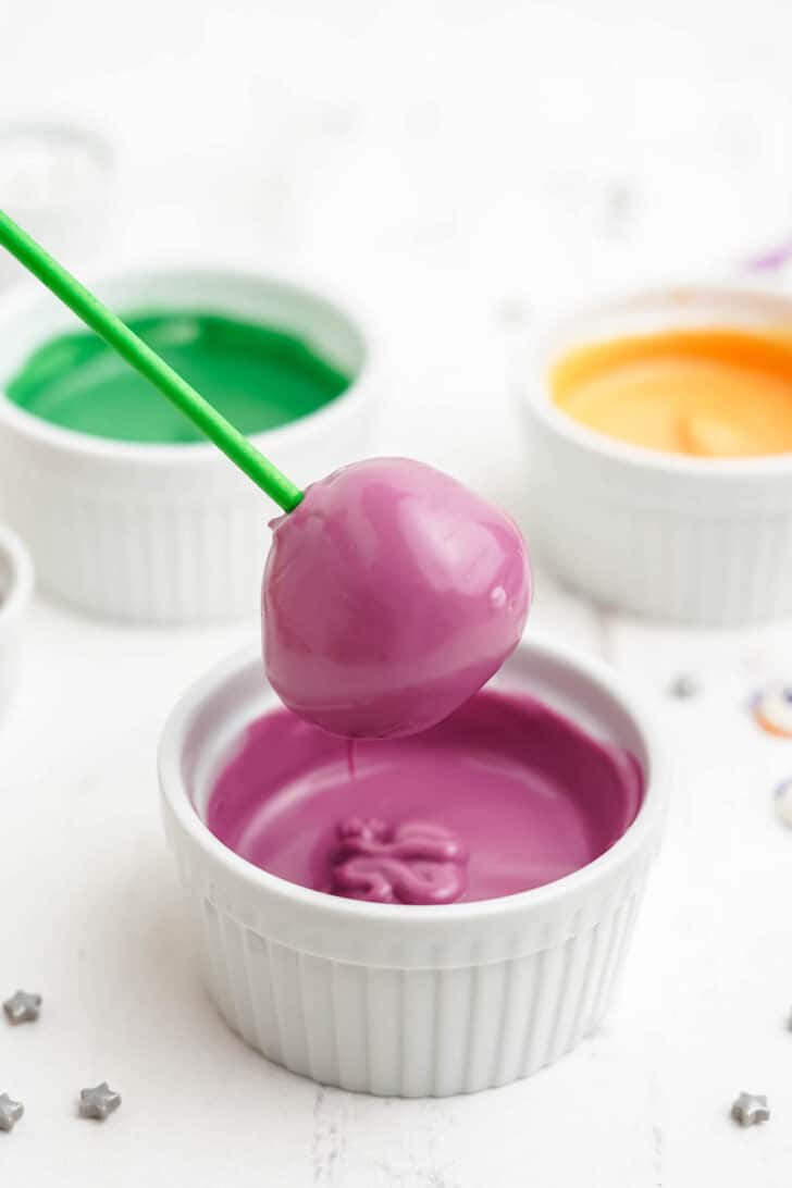 A white ramekin filled with melted purple candy coating, with a donut hole on a green lollipop stick just having been dipped into it.