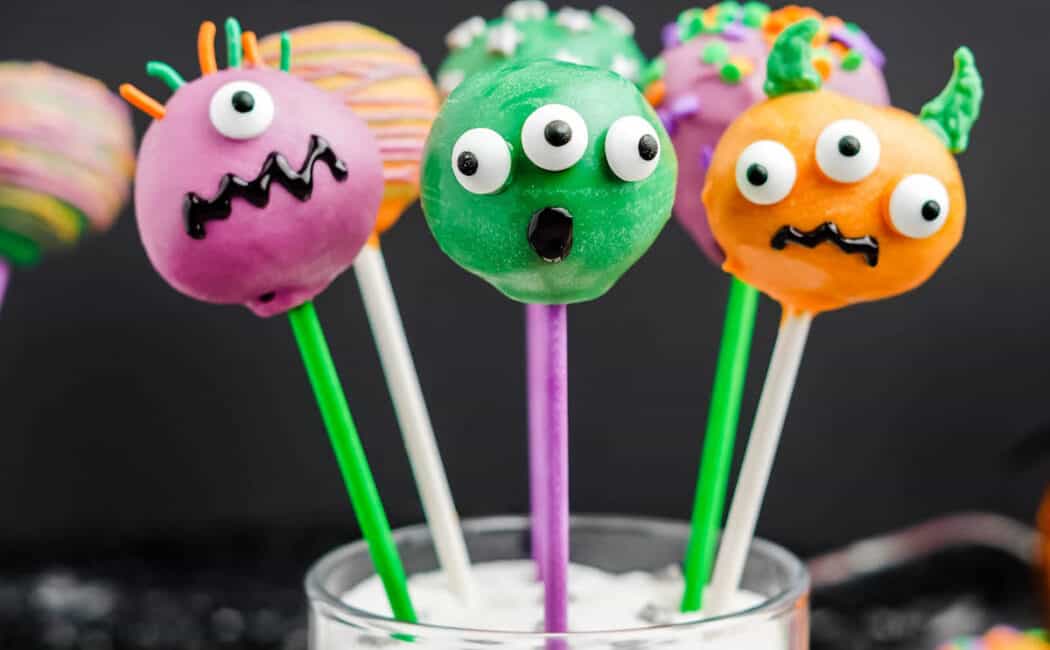 Purple, green and orange candy-coated Halloween cake pops decorated with candy eyeballs and sprinkles, sticking out a glass filled with white sugar and tied with an orange ribbon.