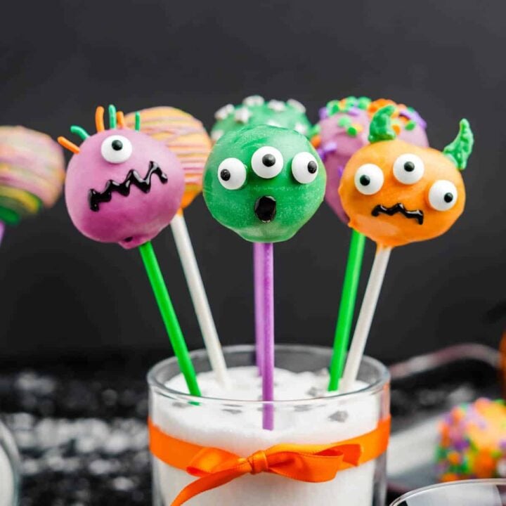 Purple, green and orange candy-coated Halloween cake pops decorated with candy eyeballs and sprinkles, sticking out a glass filled with white sugar and tied with an orange ribbon.