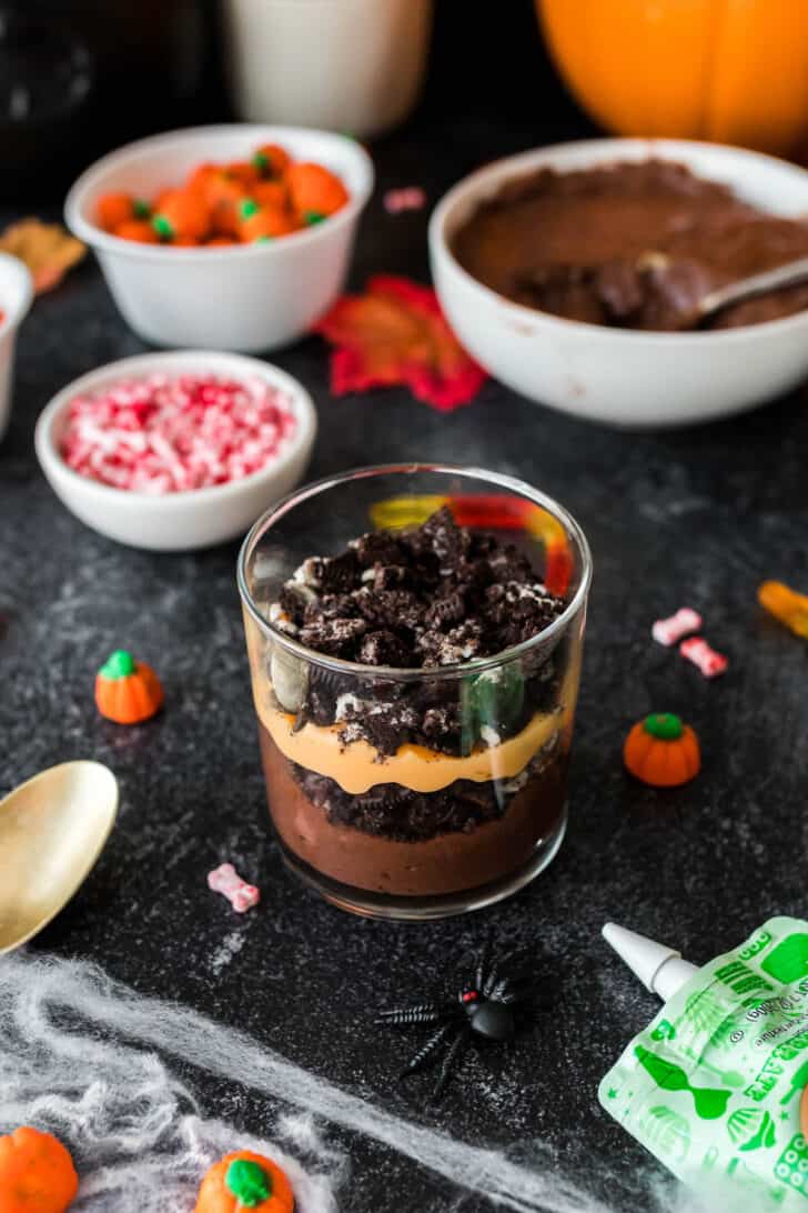 A small glass layered with Halloween dirt pudding, including layers of brown and orange puddings, crushed cookies and candy.
