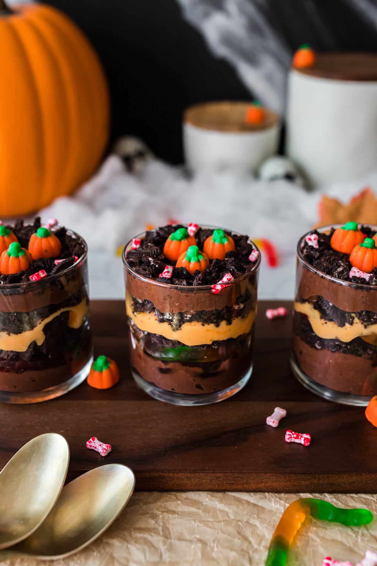 Halloween dirt cups  made with layers of chocolate and orange colored pudding, crushed cookies and gummy worms. Three cups are arranged on a wooden serving board and topped with candy pumpkins.
