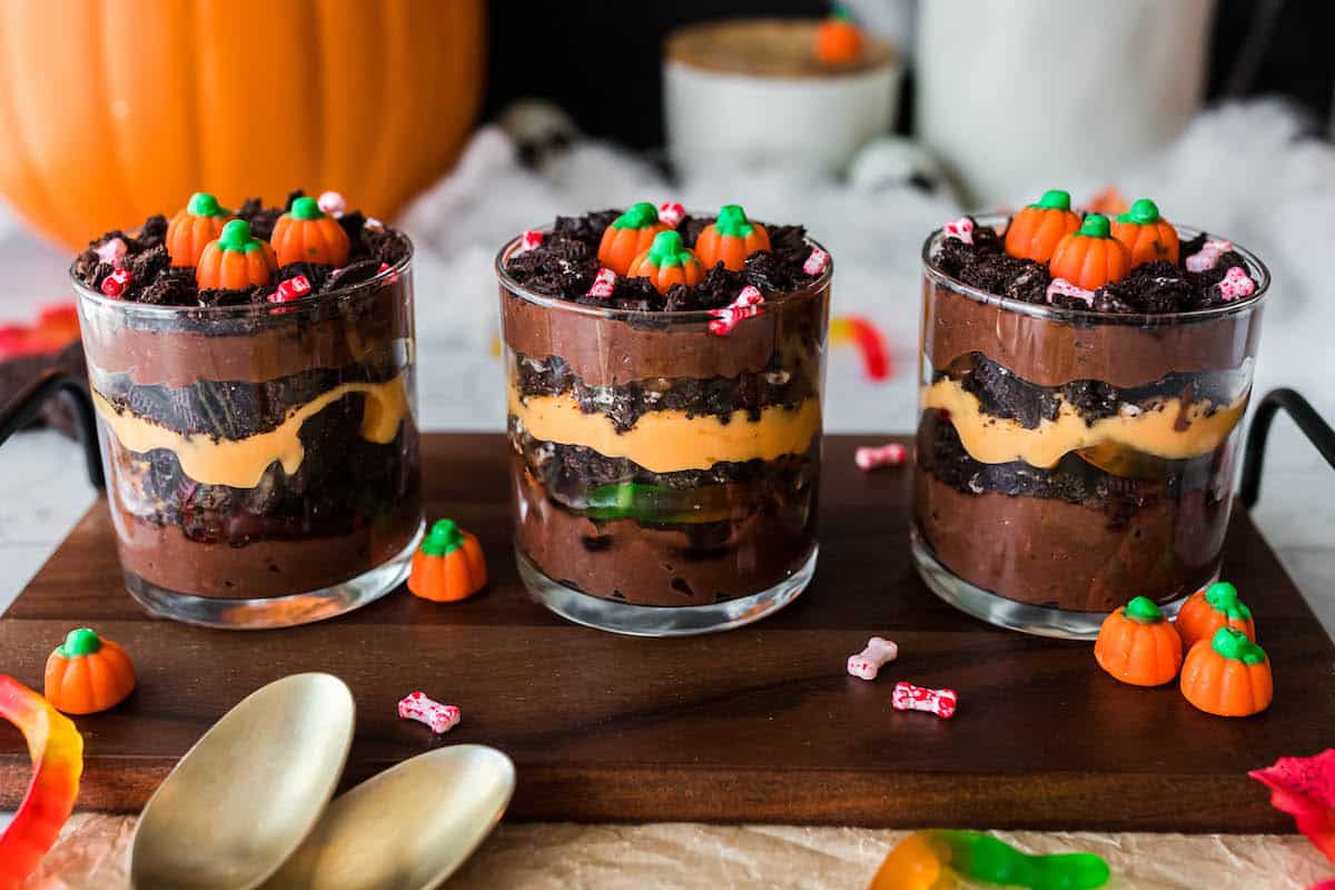 Three Halloween dirt cake cups on a wooden serving tray. Cups are made with layers of brown and orange puddings, crushed cookies and candy.