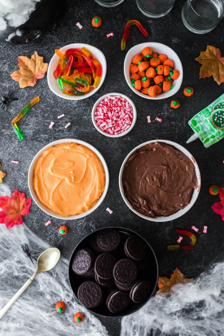 Ingredients needed for Halloween pudding cups on a dark surface, including bowls of brown and orange puddings, Oreo cookies, bone shaped candies and gummy worms.