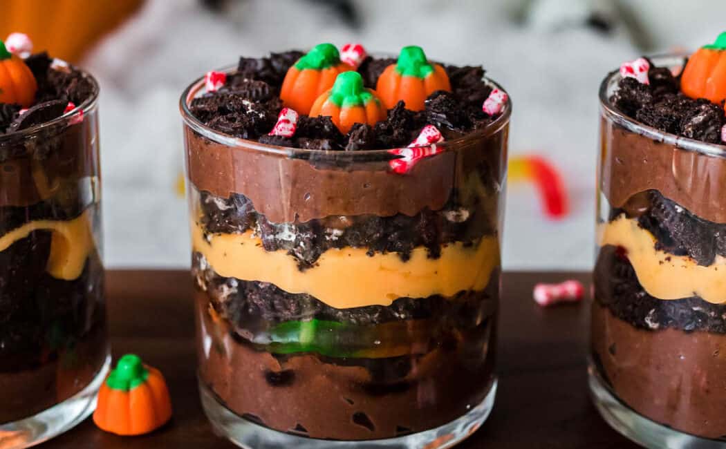 Halloween dirt cups made with layers of chocolate and orange colored pudding, crushed cookies and gummy worms. Three cups are arranged on a wooden cutting board and topped with candy pumpkins.