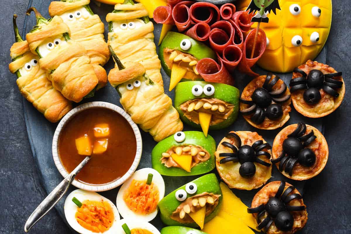 Spooky Halloween party food, like jalapeno popper mummies, apple monsters, bat tortilla chips and spider mini pizzas, arranged on a large dark platter on a dark background.