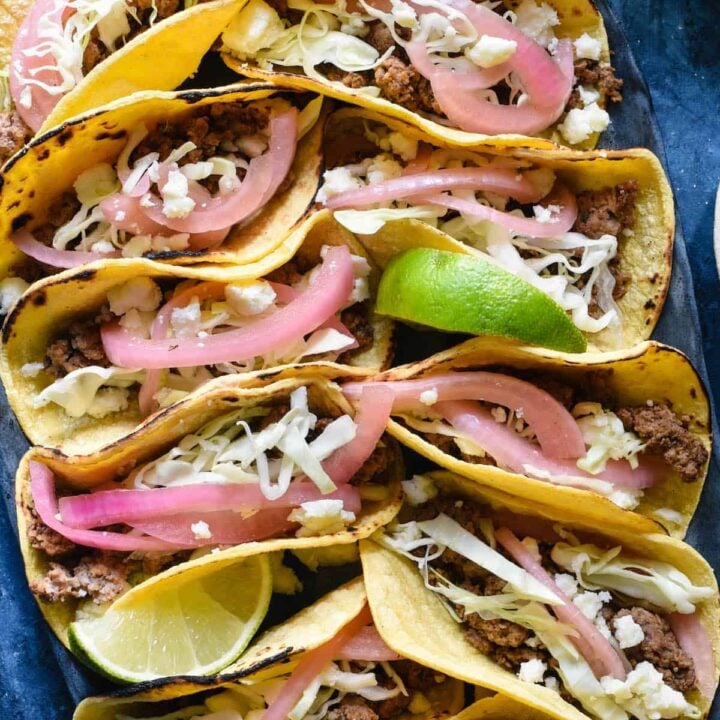 Oval tray filled with lamb tacos garnished with cheese and pickled red onions and lime wedges, on a blue surface.