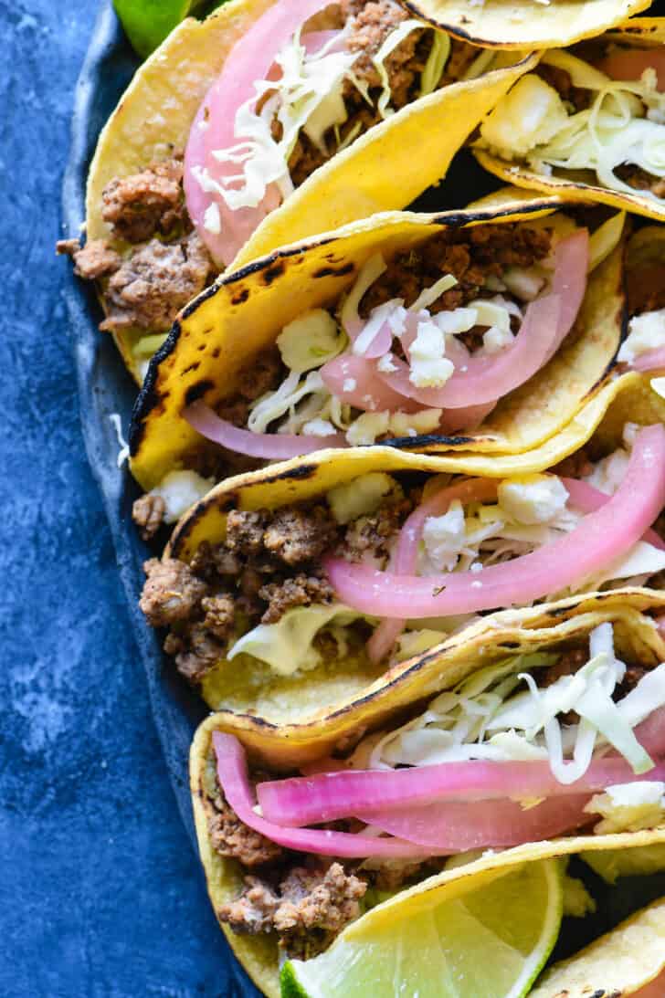 Corn tortillas filled with ground meat, topped with cabbage, pickled red onions, crumbled white cheese and lime wedges, on a blue surface.