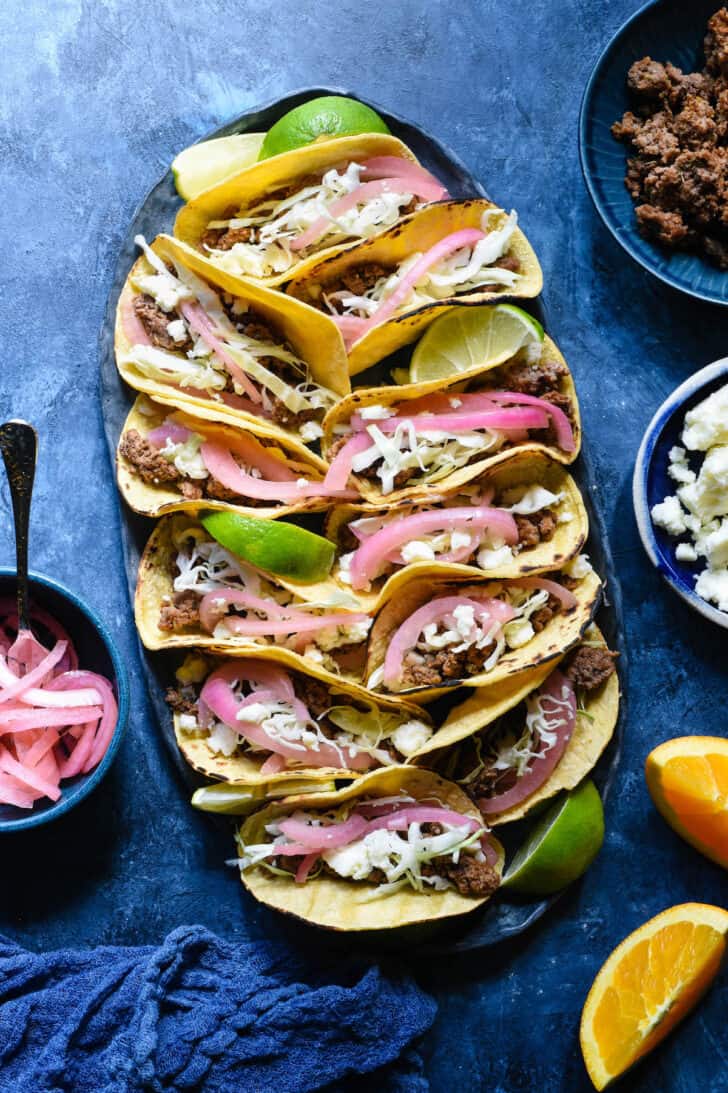 Oval tray filled with ground lamb tacos topped with cabbage, pickled red onions, crumbled white cheese and lime wedges, on a blue surface.