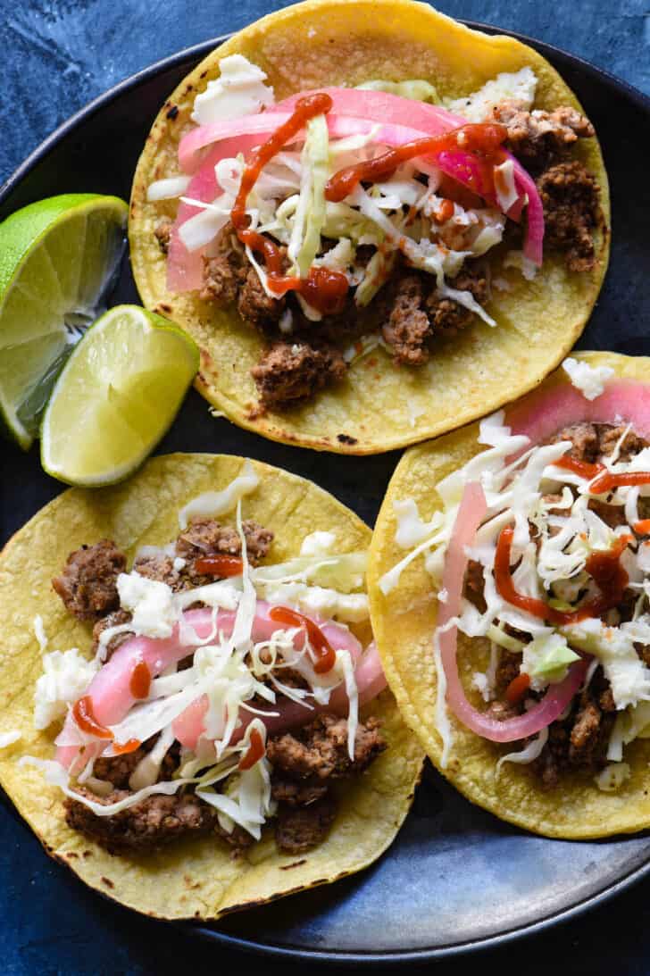 Three corn tortillas topped with ground meat, cabbage, crumbled white cheese, pickled white onions and hot sauce, on a dark plate with lime wedges.