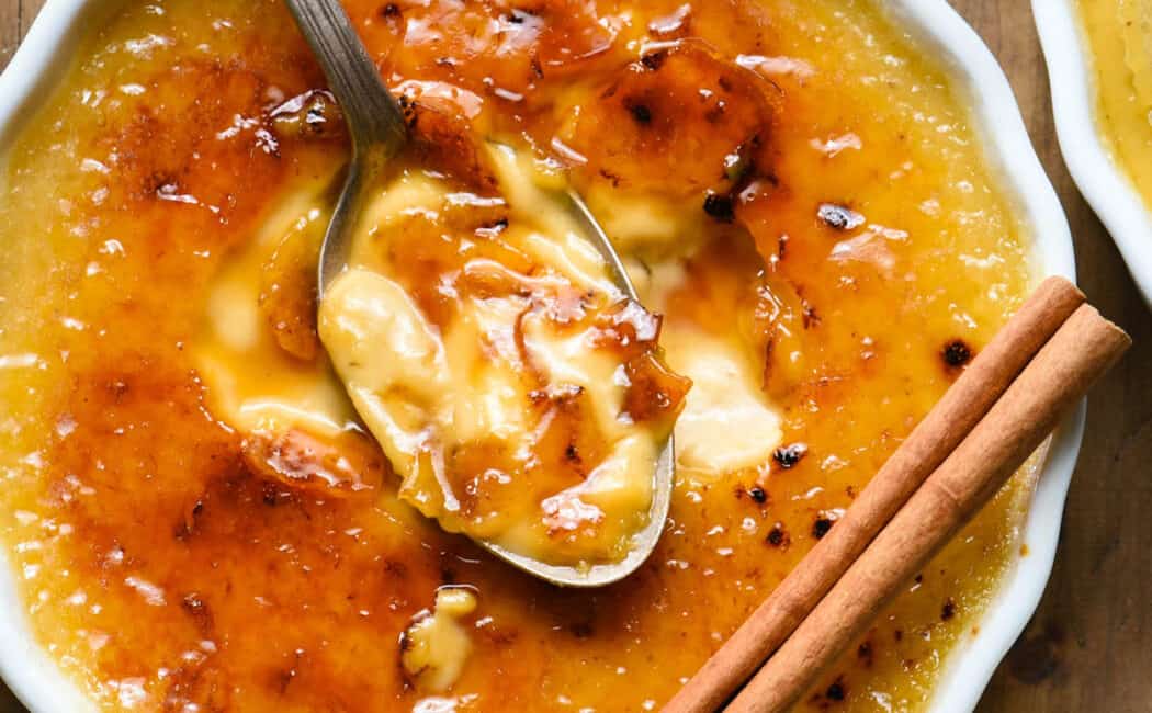 A circular white ceramic ramekin filled with an orange-hued custard topped with caramelized sugar, garnished with a cinnamon stick, with a spoon digging in.