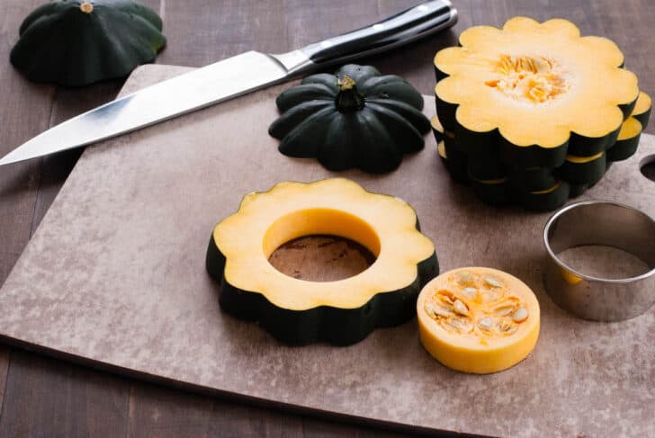 Fall gourd cut crosswise into slices, on a brown cutting board with a chef's knife and a ring mold. One gourd slice has had it's core removed with the ring mold.