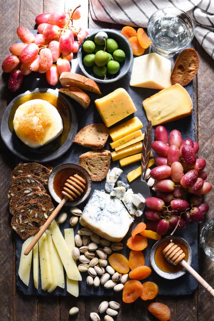 A slate cheese board filled with a variety of cheeses, pistachios, dried apricots, crackers, olives, cooked fruit and honey.