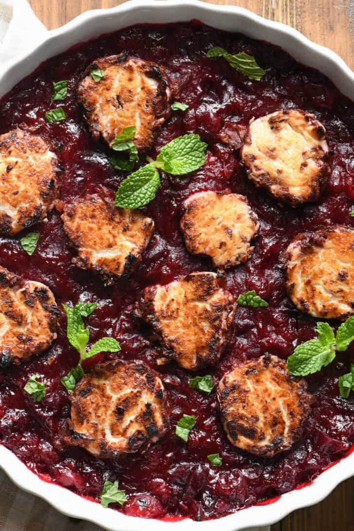 A white dish with a red berry sauce topped with baked goat cheese, garnished with fresh mint.