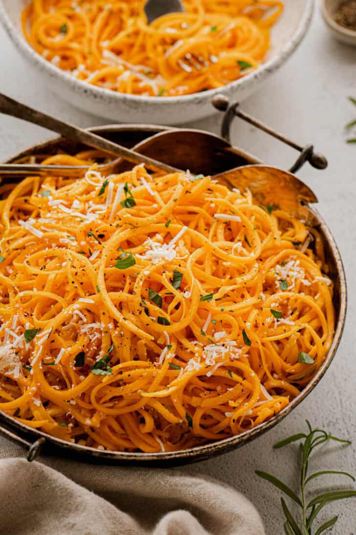 A metal bowl filled with orange gourd pasta garnished with herbs and cheese.