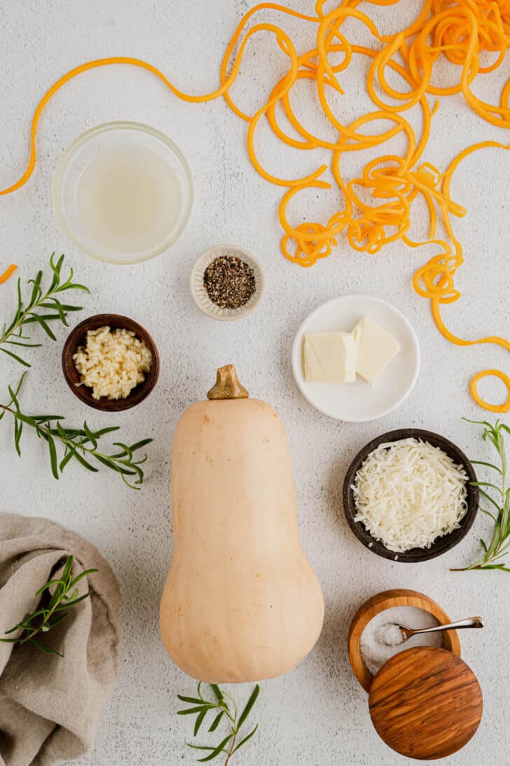 The ingredients needed for butternut squash pasta laid out on a light surface, including squash and squash noodles, butter, herbs, salt, pepper and cheese.