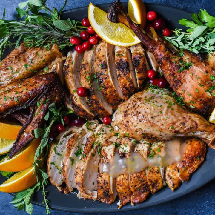 A large dark oval platter with a Cajun turkey that has been roasted and cut up arranged on it, garnished with fresh orange slices, cranberries and herb sprigs.