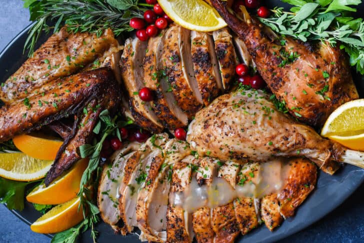 A large dark oval platter with a Cajun turkey that has been roasted and cut up arranged on it, garnished with fresh orange slices, cranberries and herb sprigs.