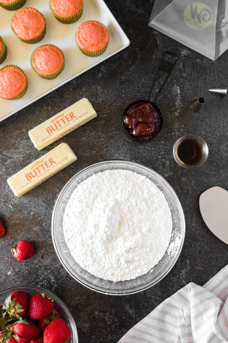 The ingredients needed for strawberry buttercream frosting laid out on a dark surface, including powdered sugar, sticks of butter, strawberry jam, and vanilla extract.