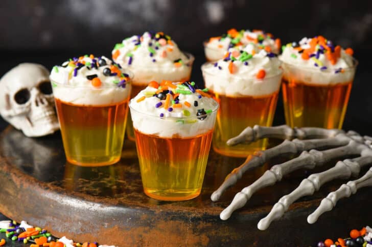 Candy corn Jello shots made with layers of yellow and orange Jello, and whipped cream, topped with sprinkles. Shots are on an upside down tray and the scene is decorated with a fake skull and skeleton hand.