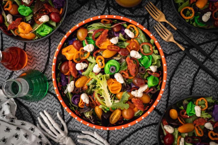 An orange bowl filled with with Halloween salad made with greens, olives, sliced peppers, dyed tortellini, mozzarella cheese skulls and pepperoni cut into bat shapes.